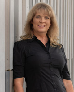 Cheryl Barton headshot, against the corrugated metal wall in the black shirt.